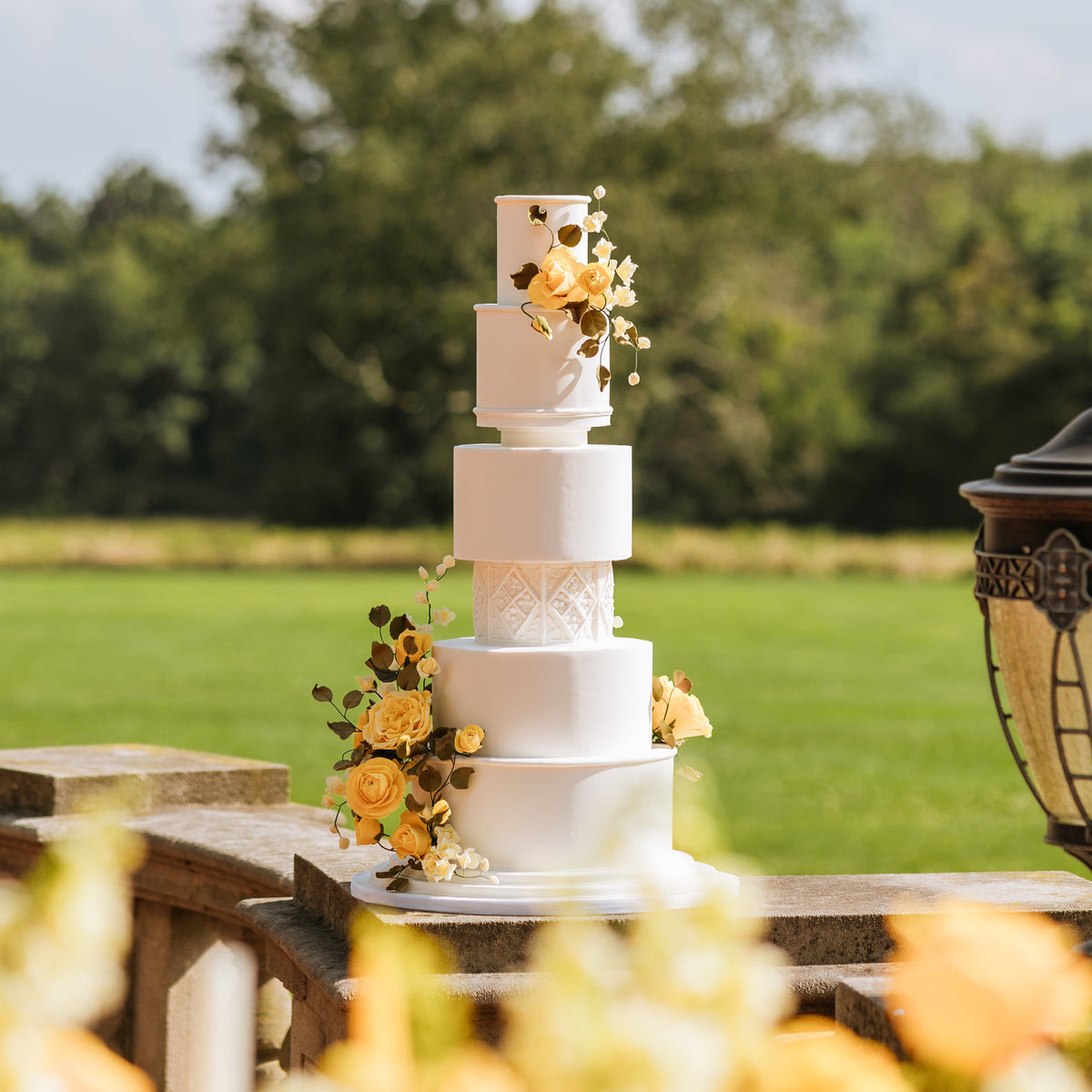 White wedding cake with yellow flowers on a table outdoors with greenery in the background