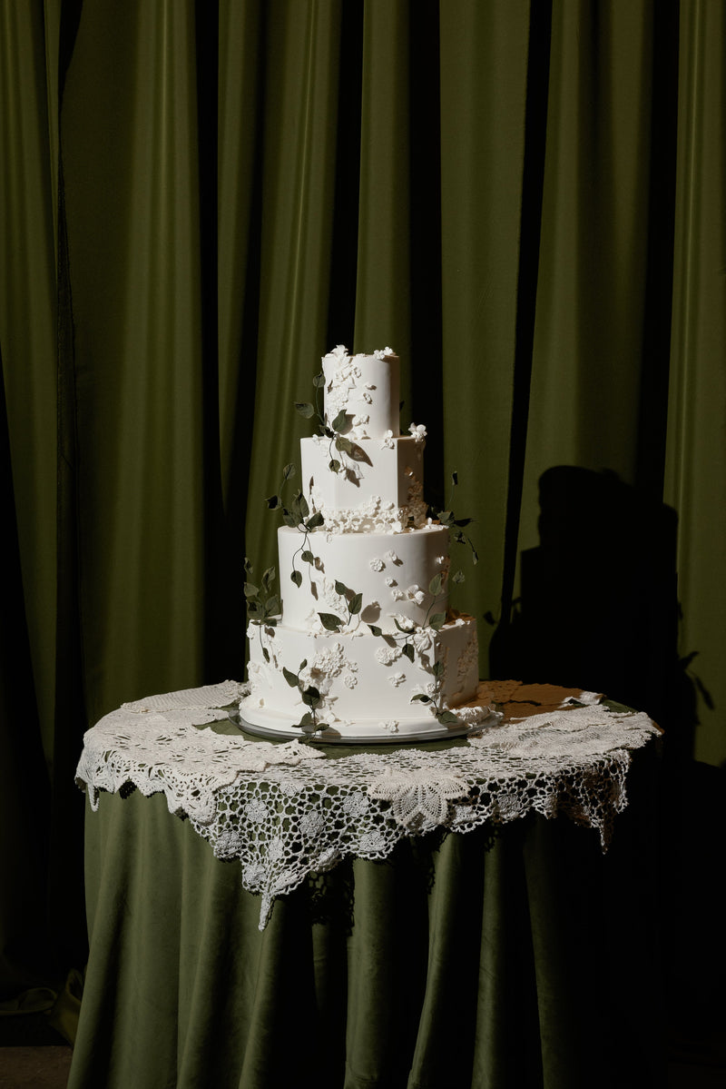 A four-tier white wedding cake decorated with dark leafy accents, displayed on a draped table with a lace doily against a dark olive green curtain backdrop.